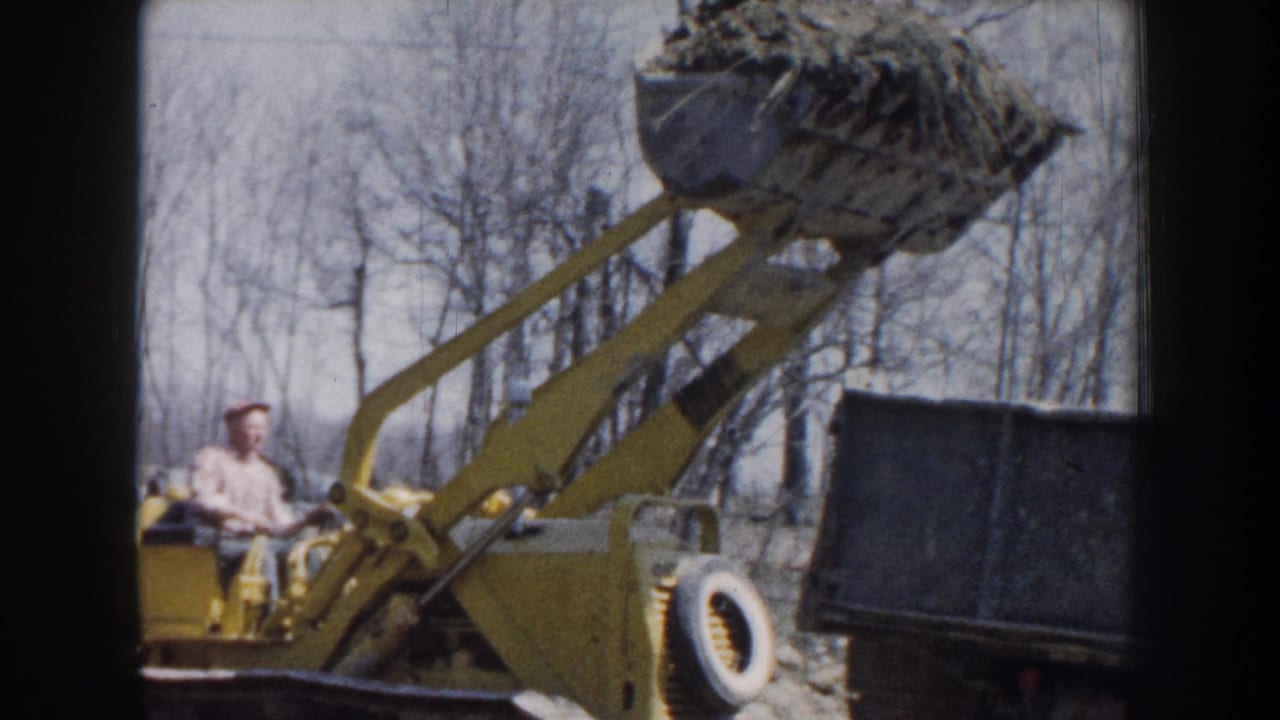 In 1959 Nebraska home-movie footage, a yellow loader lifts dirt or gravel toward a blue dump truck as a worker sits at the controls.
