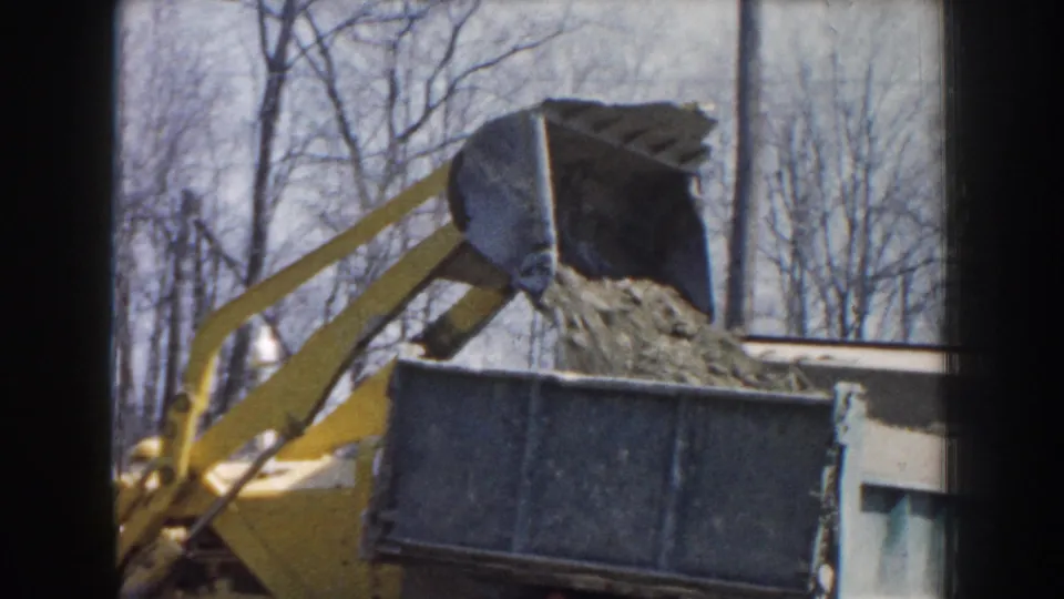 A yellow loader continues dumping dirt or gravel into the blue truck bed, framed by bare trees and soft 8mm color.