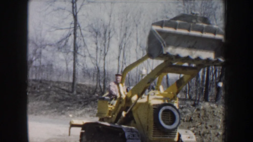 A yellow front-end loader with its bucket raised is seen near a rural roadside, with the operator partly visible and bare trees behind.