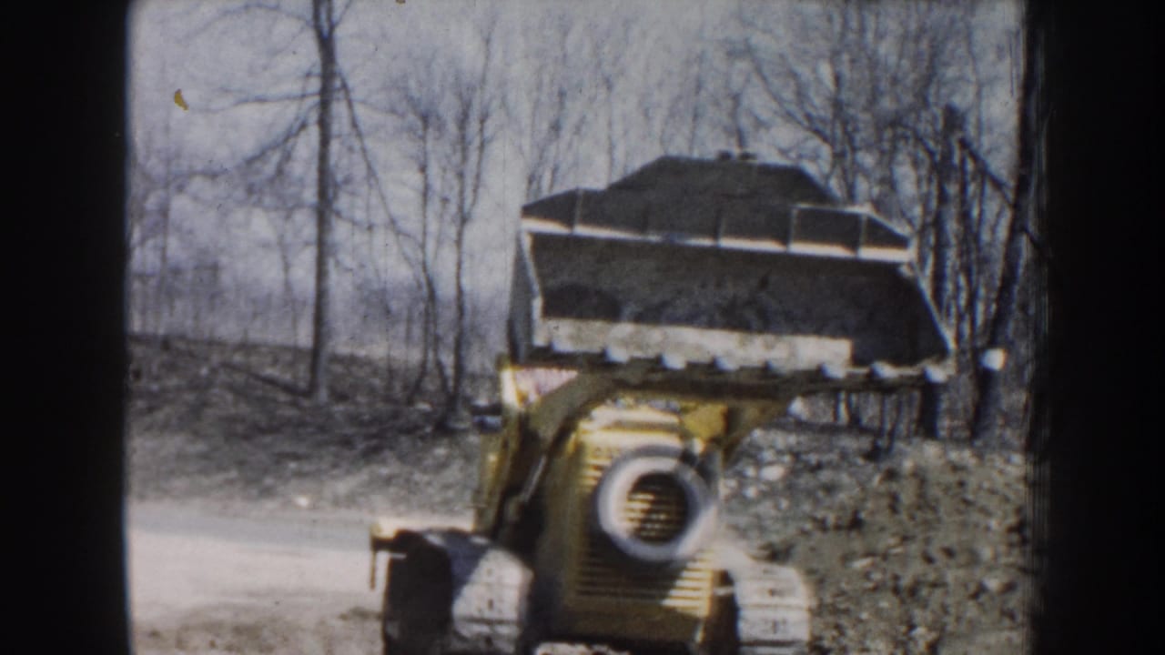 Yellow front-end loader approaches with a raised dirt-filled bucket in a wooded work area.