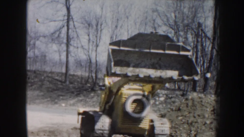 In Nebraska 1959 home-movie footage, a yellow front-end loader moves toward camera with a raised bucket of dirt near bare trees.