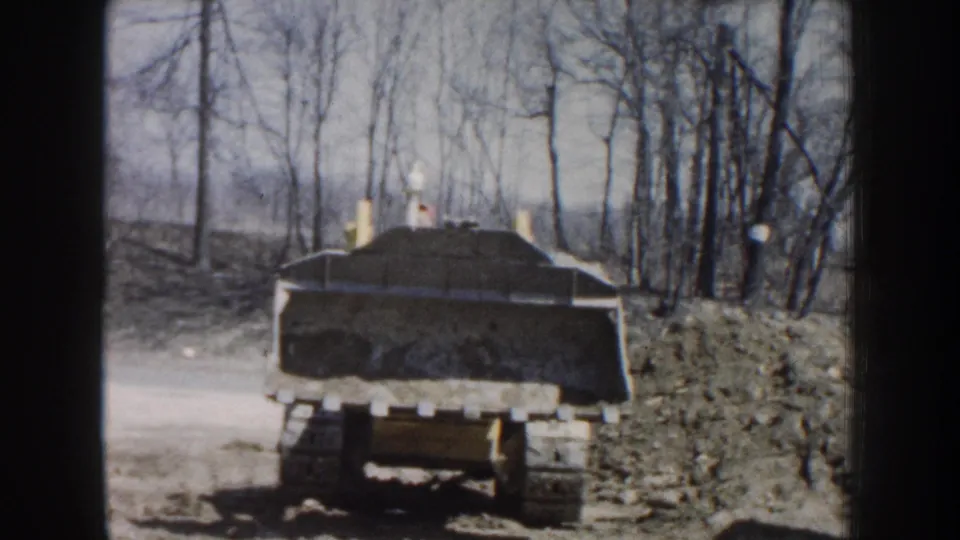 The loader is partly turned in a wooded Nebraska work area, with the bucket raised and the operator only faintly visible.
