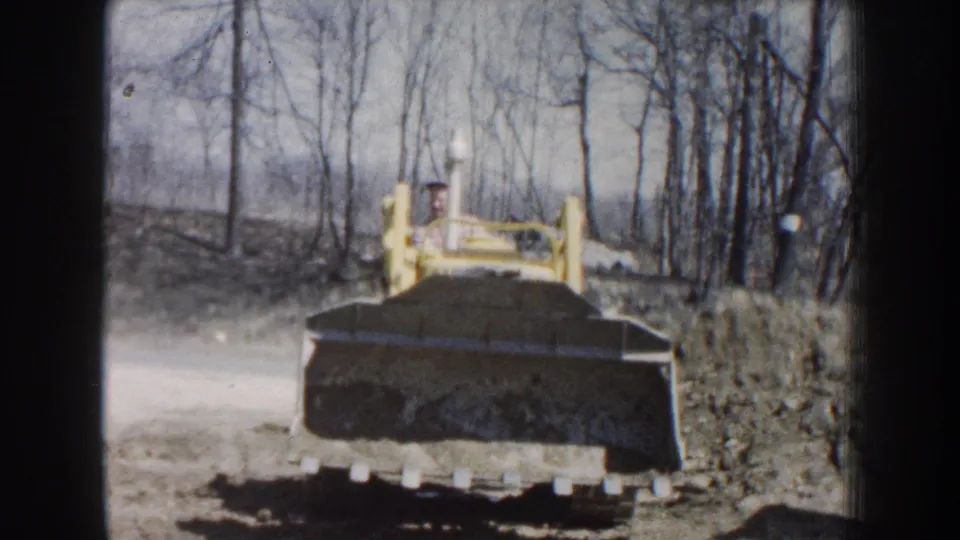 A yellow front-end loader faces camera in 1959 Nebraska footage, its bucket filled with soil against a backdrop of bare trees.