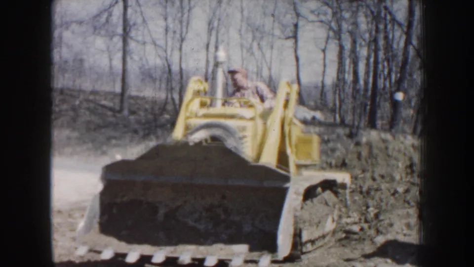 A man in a light shirt and cap operates the yellow loader as its filled bucket passes beside a dirt bank in Nebraska.
