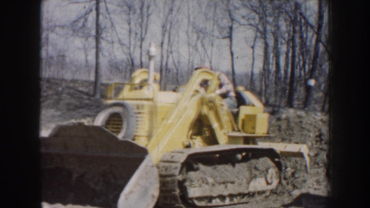 1959 Nebraska 8mm frame showing a yellow tracked bulldozer with an adult male operator working beside bare trees.