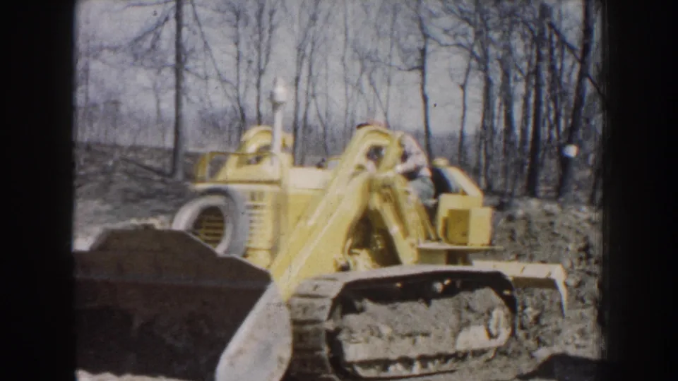 1959 Nebraska 8mm frame showing a yellow tracked bulldozer with an adult male operator working beside bare trees.