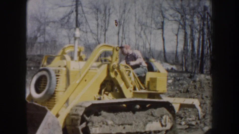 1959 Nebraska home-movie frame of a man seated on a yellow tracked bulldozer with bare trees in the background.