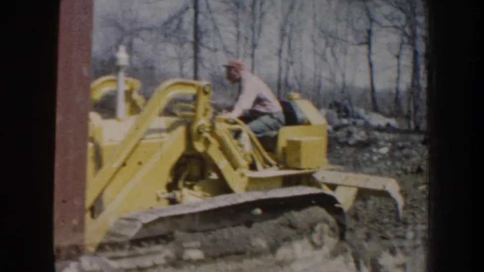 1959 Nebraska frame showing an adult man in work clothes driving a yellow tracked bulldozer through a wooded dirt area.