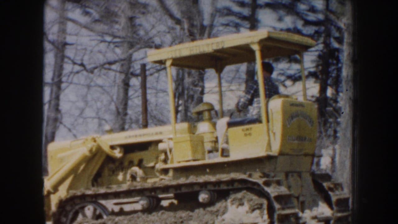 In Nebraska 1959 footage, a man sits under the canopy of a yellow tracked bulldozer beside leafless trees and uneven dirt.