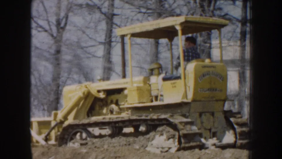 The 1959 Nebraska home-movie frame shows a yellow Caterpillar-style crawler tractor with a man driving across rough dirt.