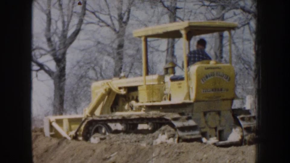 A man in a plaid shirt operates the yellow bulldozer on a dirt rise, with leafless trees in the Nebraska background.