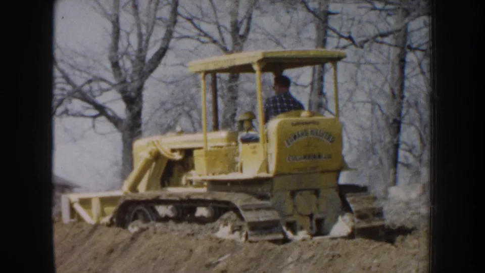 The frame shows the rear and side of a yellow bulldozer with visible branding as a man operates it in Nebraska, 1959.