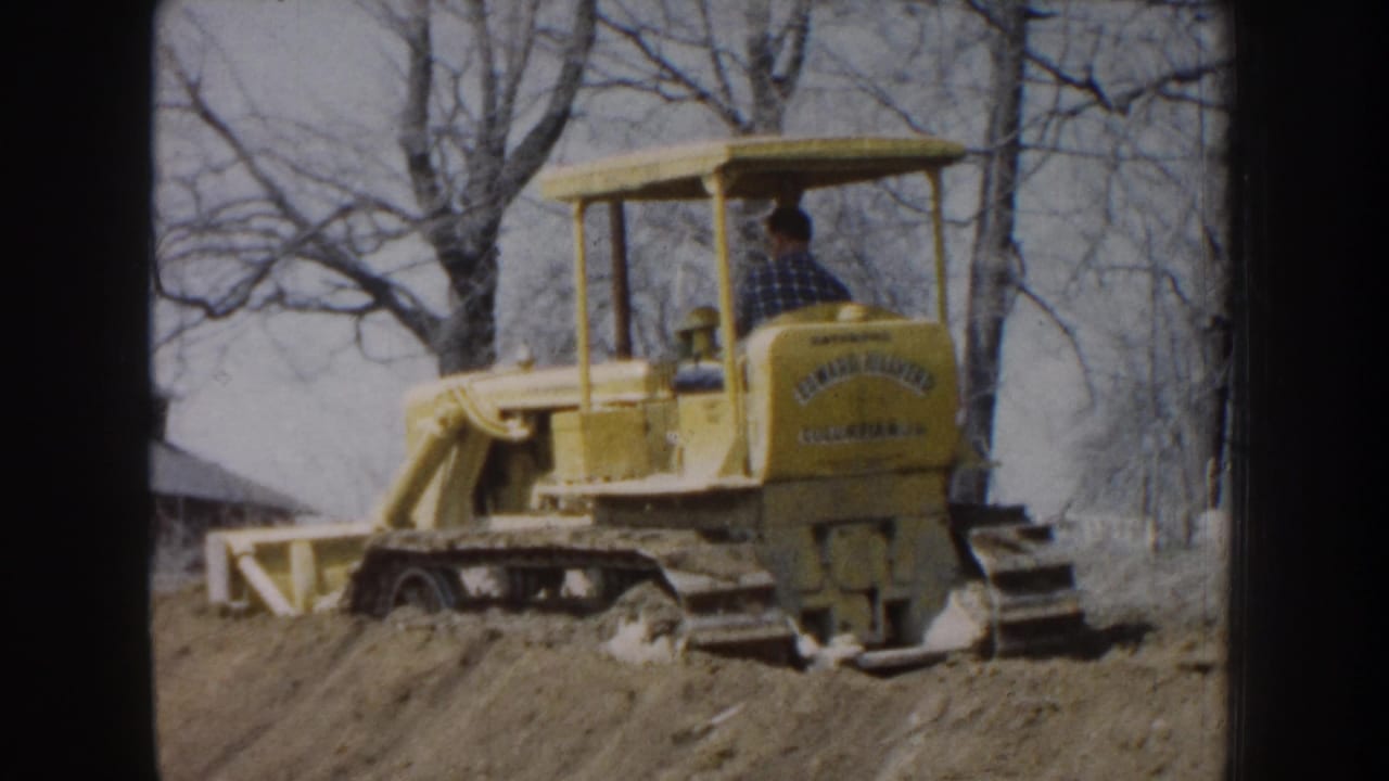1959 Nebraska 8mm frame showing a yellow tracked bulldozer moving dirt near bare trees with an operator seated at the controls.