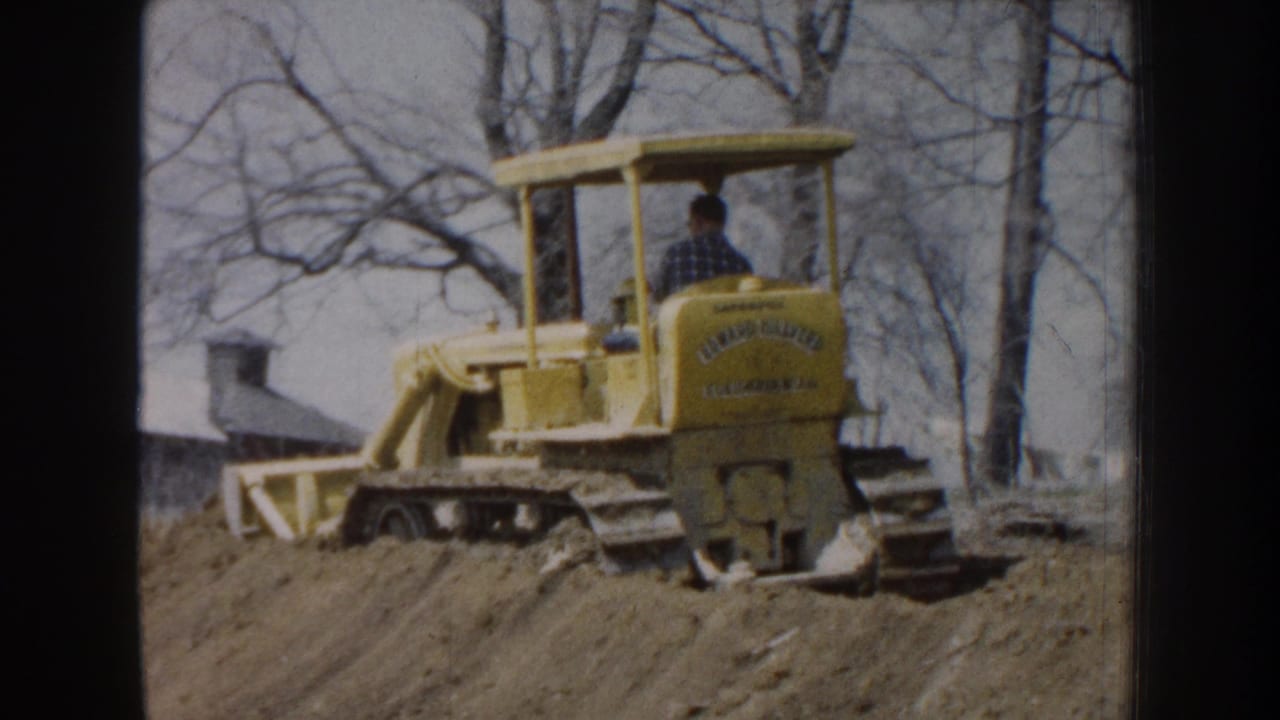 1959 Nebraska rural construction frame with a yellow bulldozer, seated operator, dirt mound, bare trees, and a distant farm-like structure.