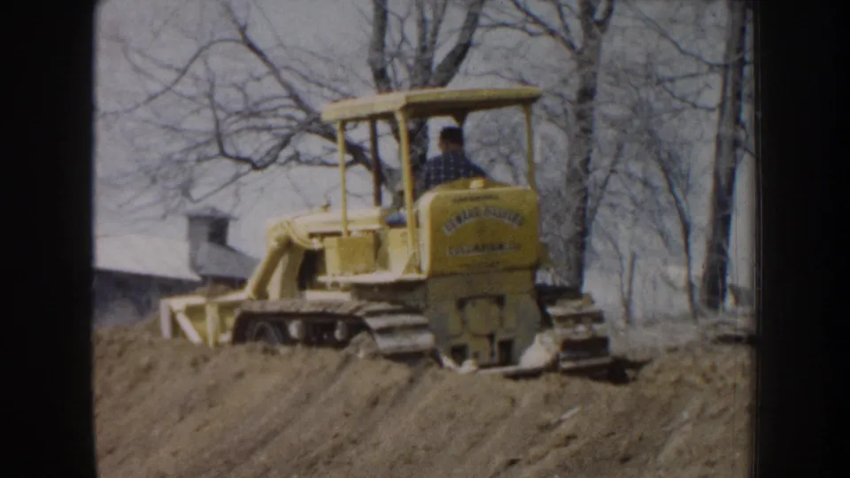 1959 Nebraska 8mm still of a yellow tracked bulldozer working dirt, with the operator seen from behind against leafless trees.
