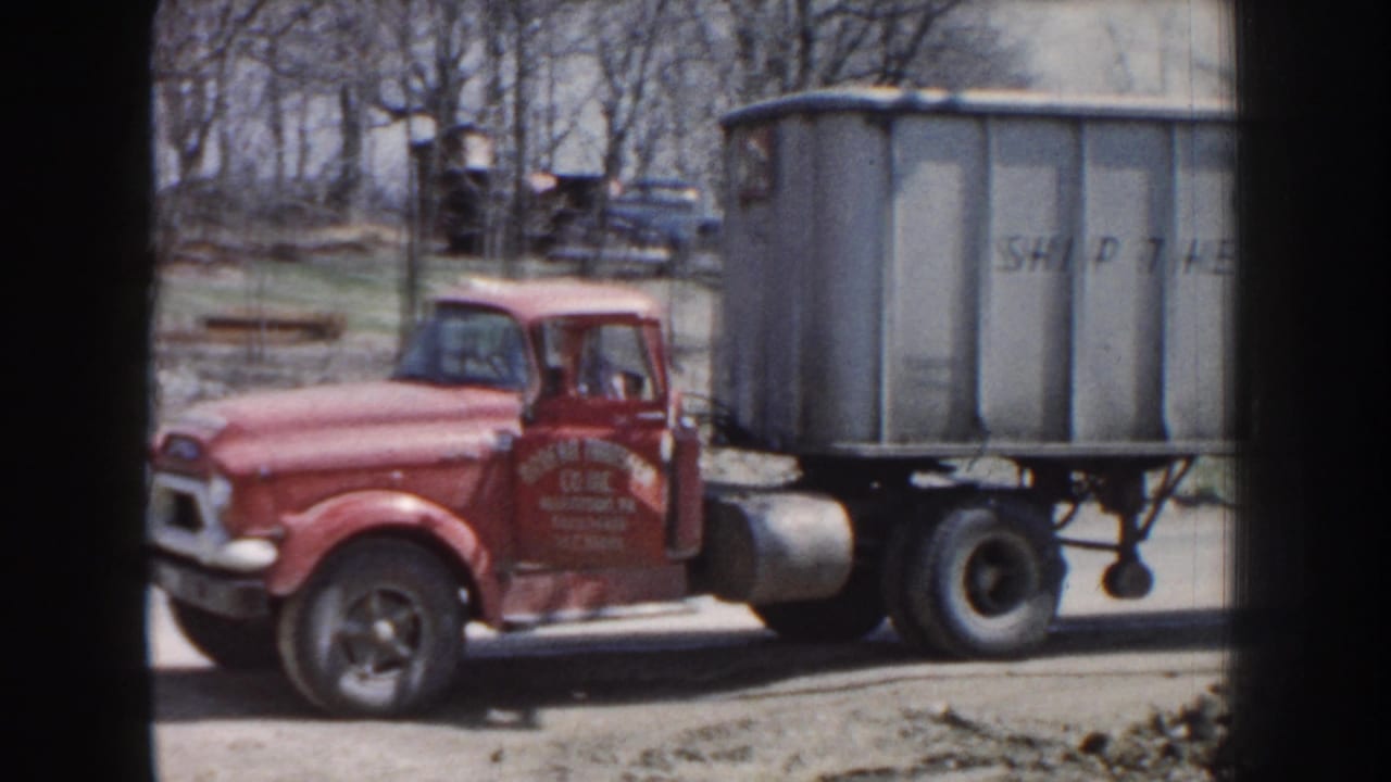 Red truck cab with a driver visible next to a gray trailer marked with partial lettering.