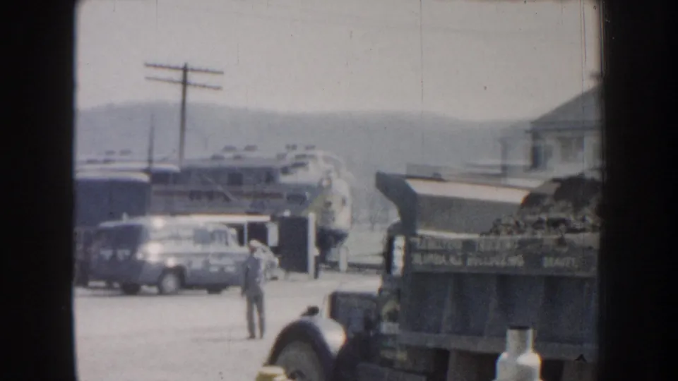 A silver passenger train approaches a Nebraska yard area in 1959, with a van, suited man, utility pole, and buildings nearby.