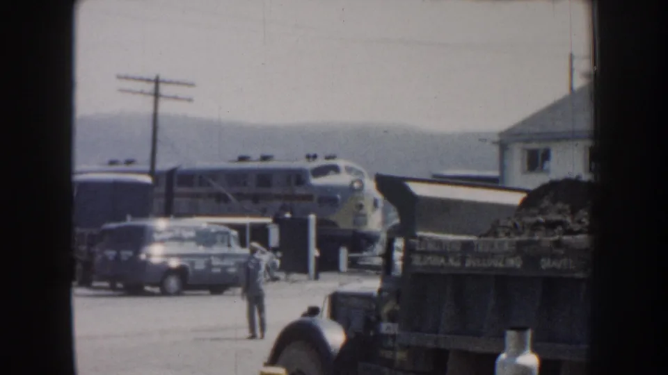 The train yard scene holds on the approaching silver passenger train, with a man standing near a vintage van and station-side buildings.