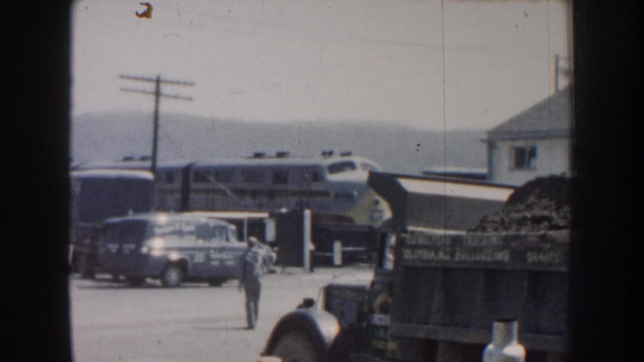 1959 Nebraska home-movie frame showing a passenger train beside cars, a dump truck, utility poles, and a man crossing the station lot.