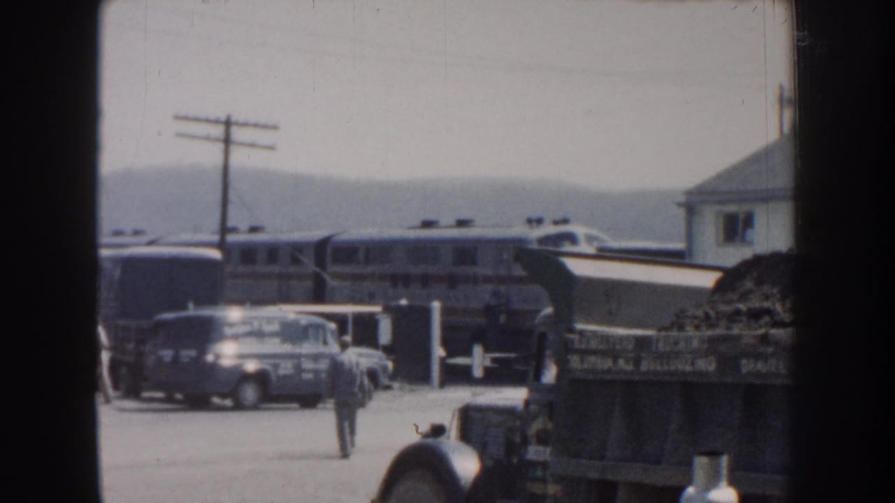 1959 Nebraska rail-station view with a passenger train, parked cars, a dump truck, utility poles, and a man seen from a distance.