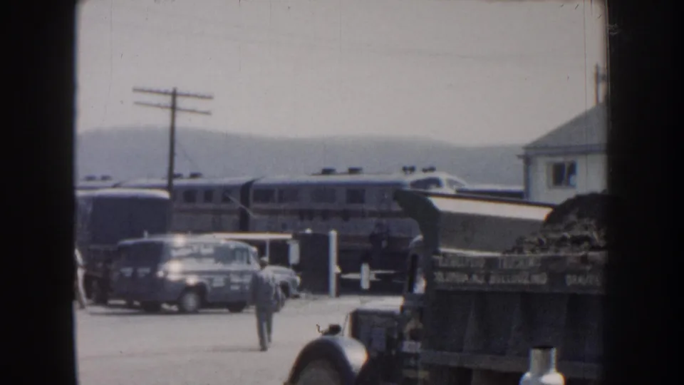 1959 Nebraska rail-station view with a passenger train, parked cars, a dump truck, utility poles, and a man seen from a distance.