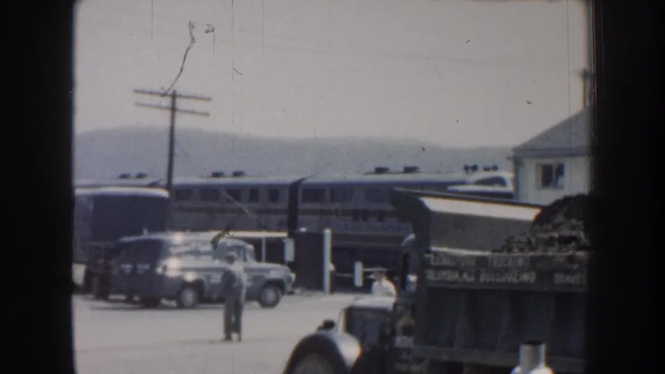 1959 Nebraska station-lot frame with a man near cars, a passenger train, a dump truck, utility poles, and hazy hills in the background.