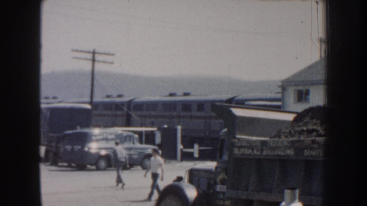 Men cross an industrial yard with vehicles and a train in the background.