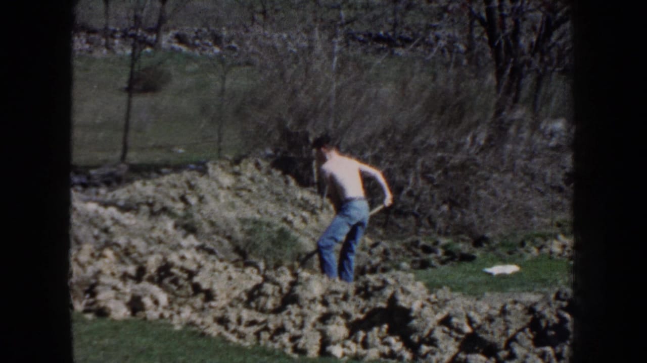 1959 Nebraska 8mm frame of a shirtless man working with a shovel in rocky ground near grass, brush, and leafless trees.