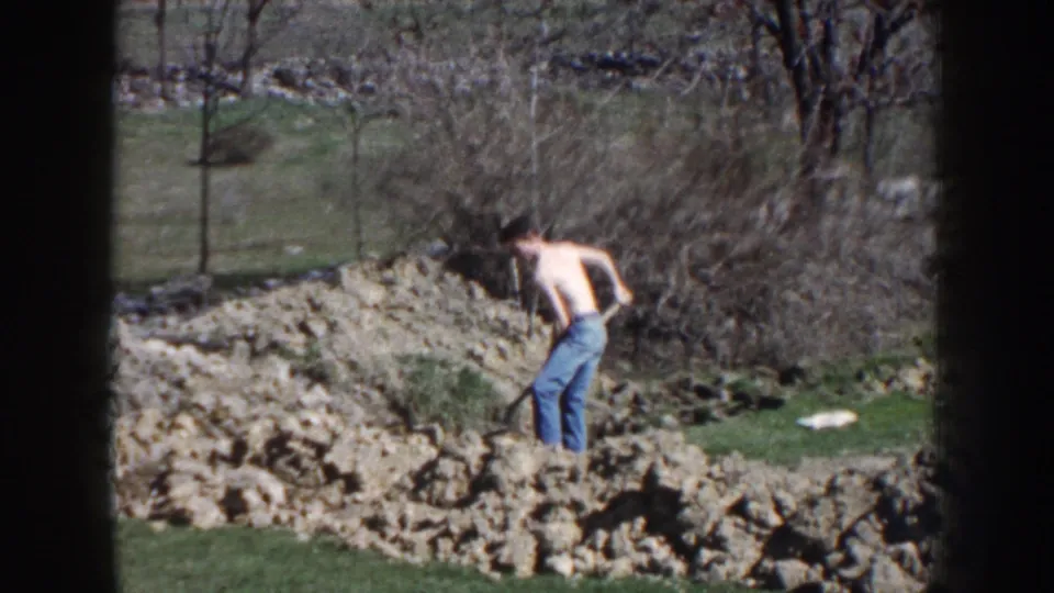 1959 Nebraska 8mm frame showing a man pausing with a shovel amid rocky soil, grass, brush, and bare trees outdoors.