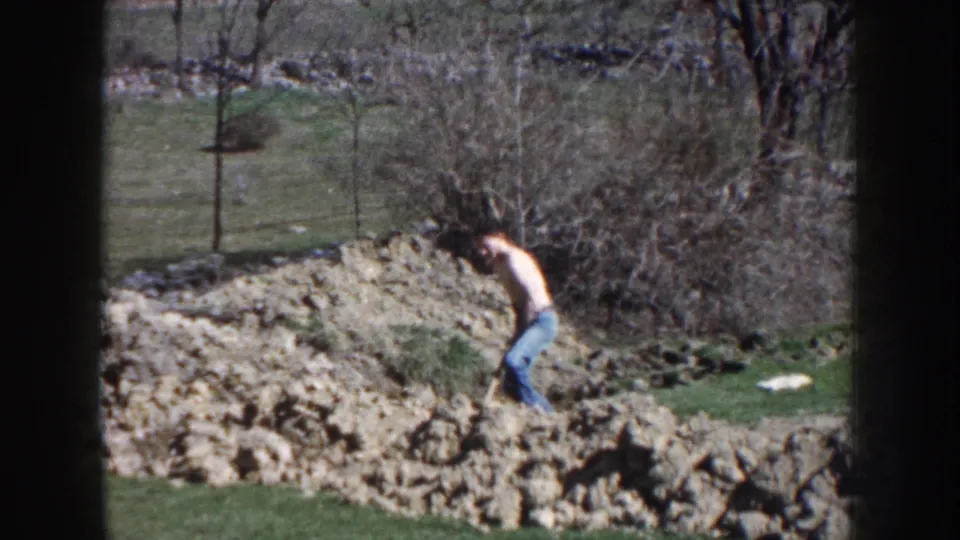 1959 Nebraska 8mm frame of a man bending into shoveling work on rocky ground with grass and bare trees behind him.