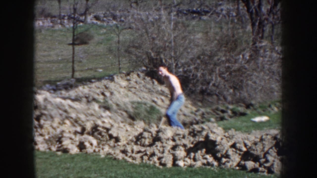In Nebraska 1959 home-movie footage, a shirtless man in blue jeans works with a shovel beside turned soil in a grassy yard.