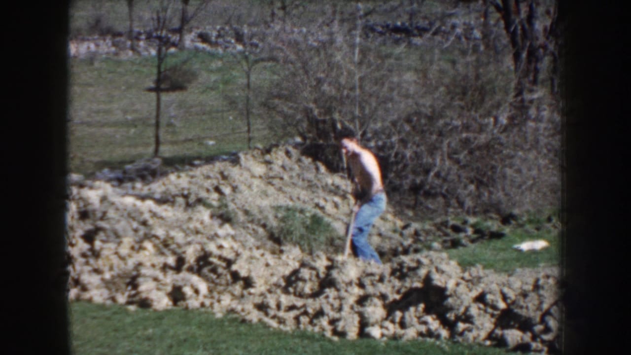 The shirtless man leans into the shovel near loose earth, with grass, bare trees, and a fence visible behind him.