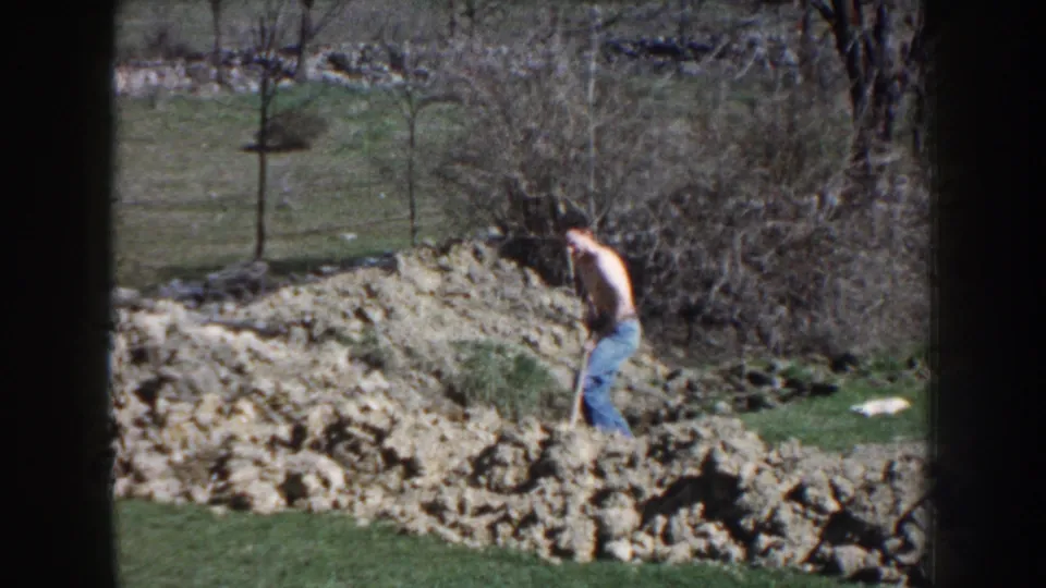 The shirtless man leans into the shovel near loose earth, with grass, bare trees, and a fence visible behind him.