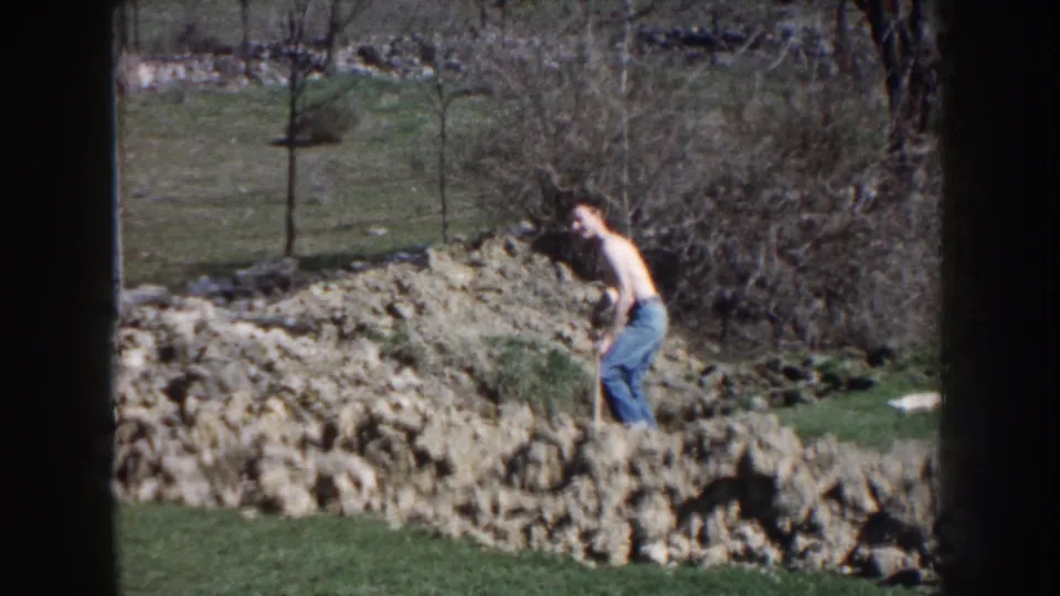 A distant 8mm frame shows the man beside a shovel and uneven soil in a sunlit Nebraska yard setting.
