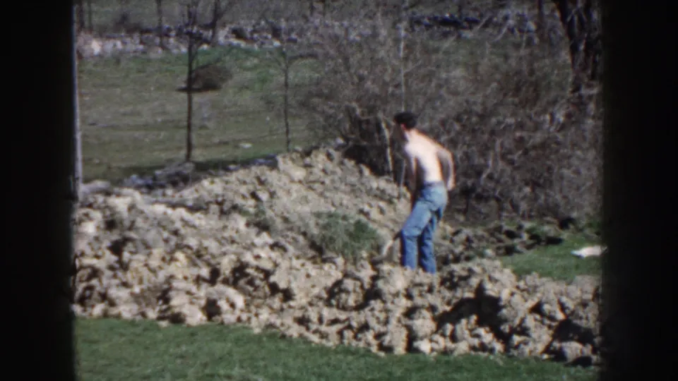 The man faces the soil mound with the shovel upright, framed by grass, brush, bare trees, and daylight in the rural yard.