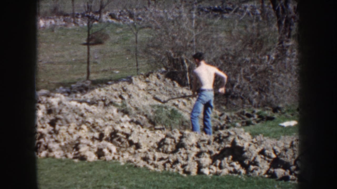 In 1959 Nebraska home-movie footage, a shirtless man in blue jeans works with a shovel beside turned earth in a grassy rural area.