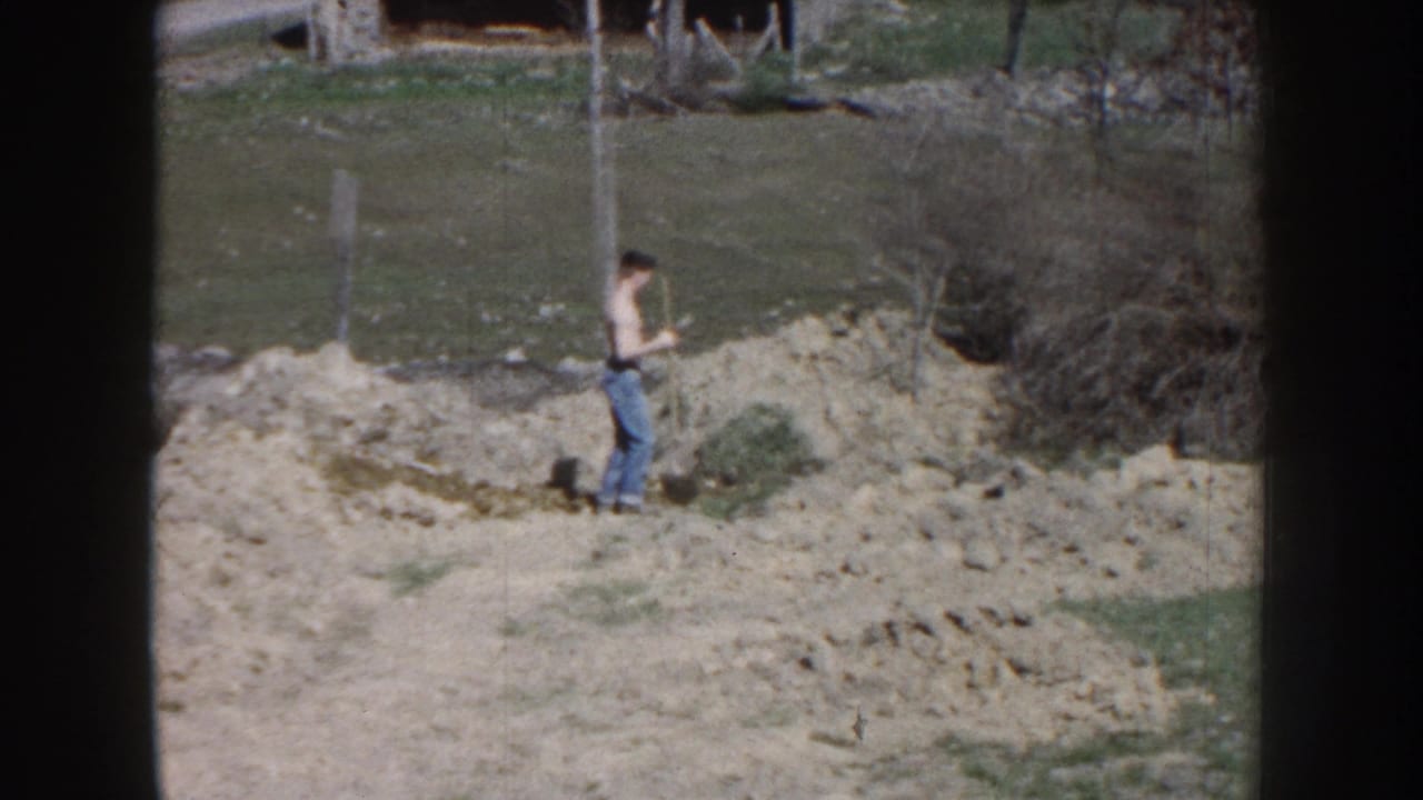 Man stands with a shovel near dirt piles and a fence post.