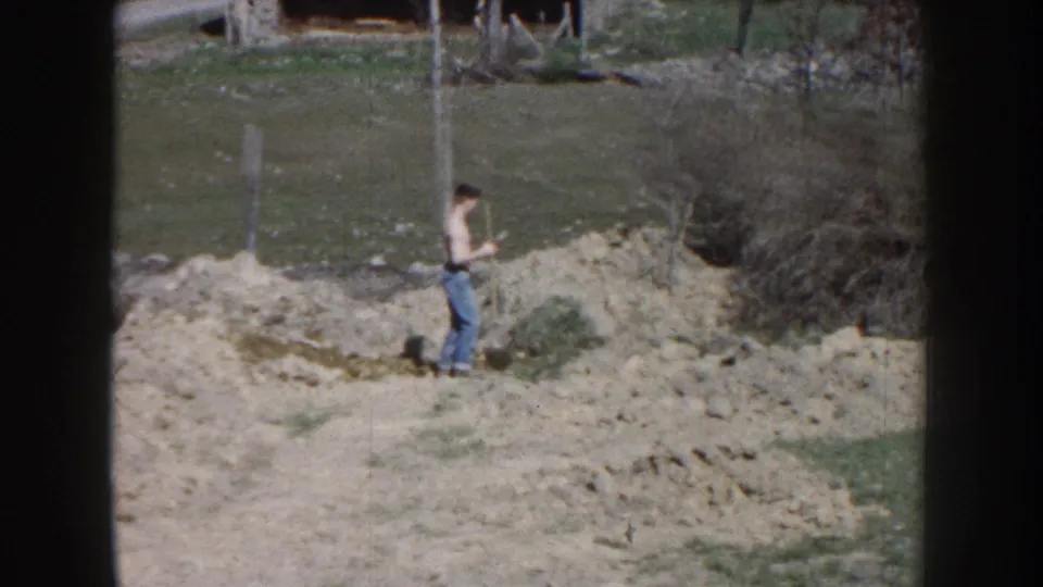 The man pauses or steps across the work area, with pale dirt mounds, grass, bare brush, and fence posts behind him.