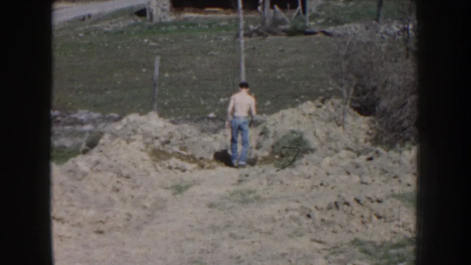 A distant view shows the shirtless man from behind as he works near the central dirt pile in the rural Nebraska field.