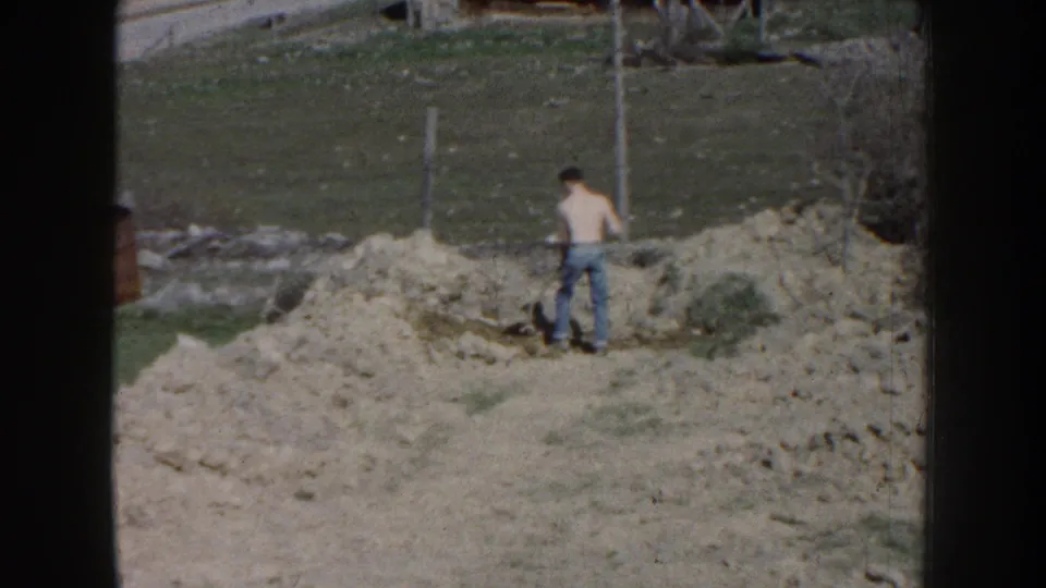 The final frame holds on the man near the disturbed soil, with patchy grass, bare vegetation, and fence posts in the background.
