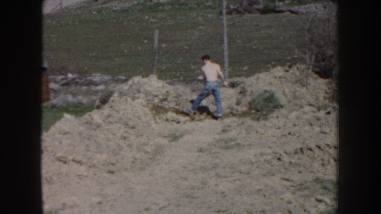 Shirtless man in jeans walking with a shovel near dirt piles in a grassy yard.