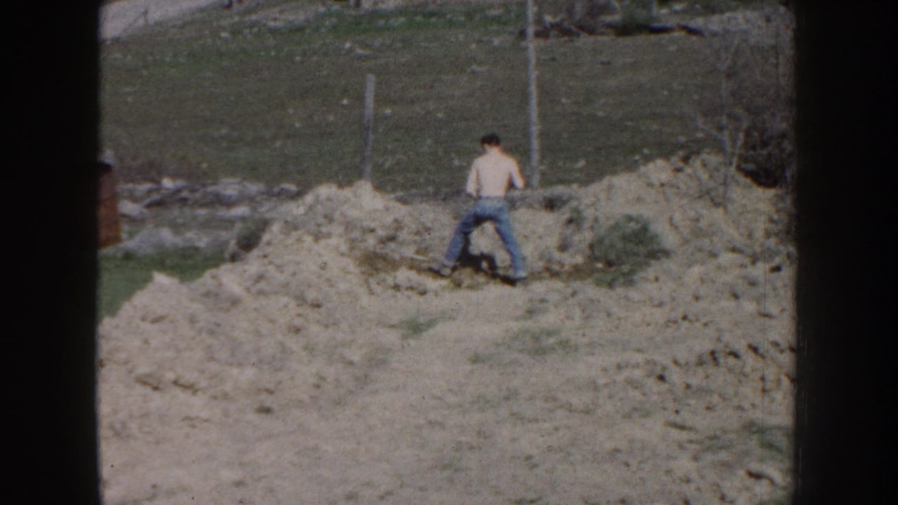 Man in jeans stands with a shovel beside a long mound of dirt outdoors.
