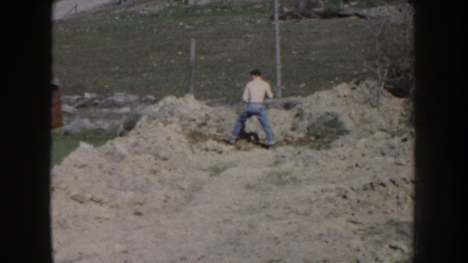A shirtless man appears to work with a shovel beside a dirt mound in a rural Nebraska yard in 1959 footage.