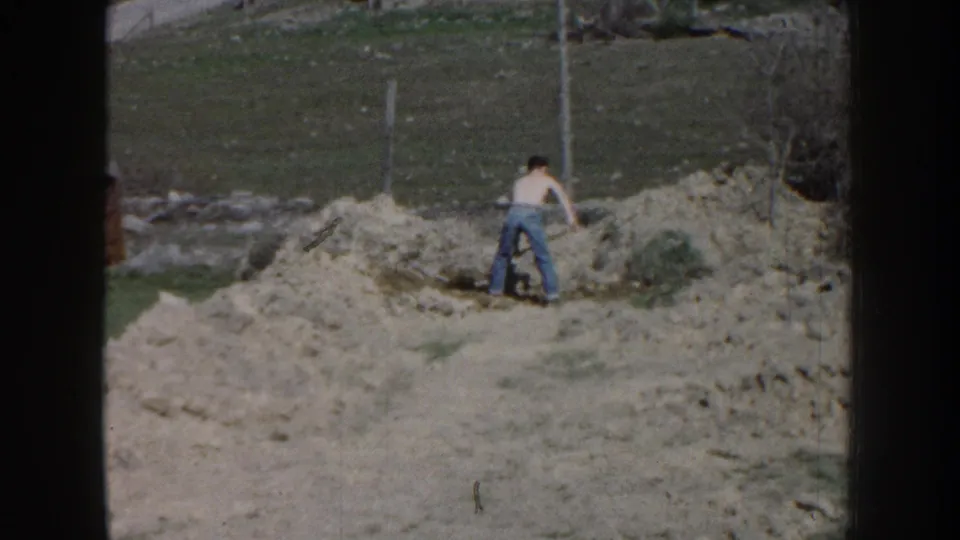 The 1959 Nebraska footage shows the man bending toward the soil as he digs or moves dirt in the yard.