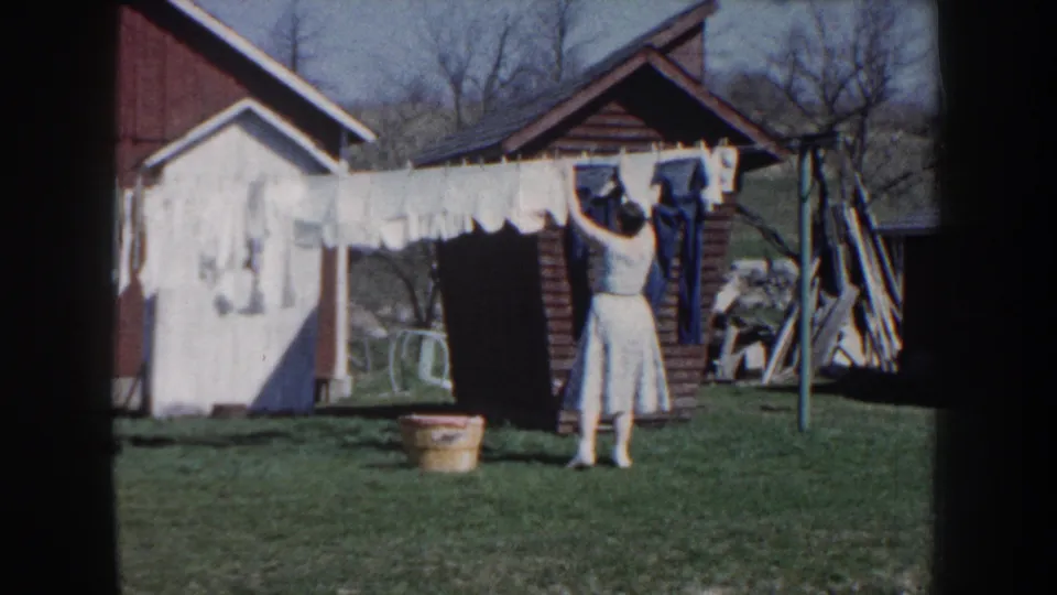 In a rural Nebraska yard, a woman in a light dress hangs clothing on a clothesline between small outbuildings.
