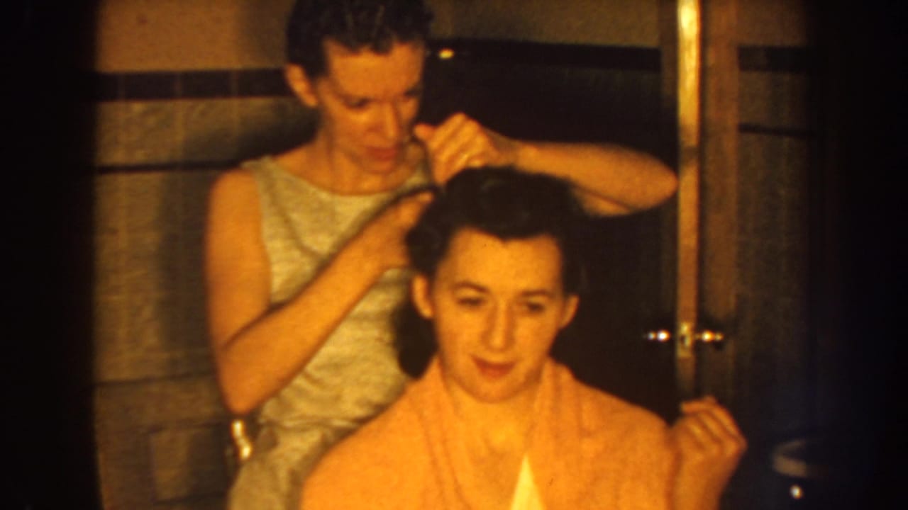 Standing woman combs a seated woman's dark hair in a tiled bathroom.