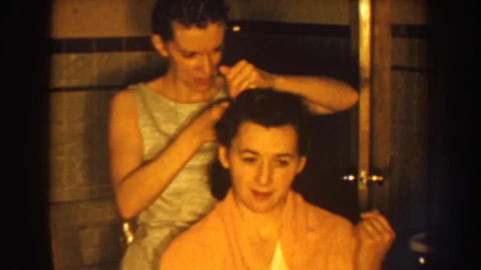 In 1959 Nebraska home-movie footage, a standing woman combs a seated woman's dark hair as she smiles in a tiled bathroom.