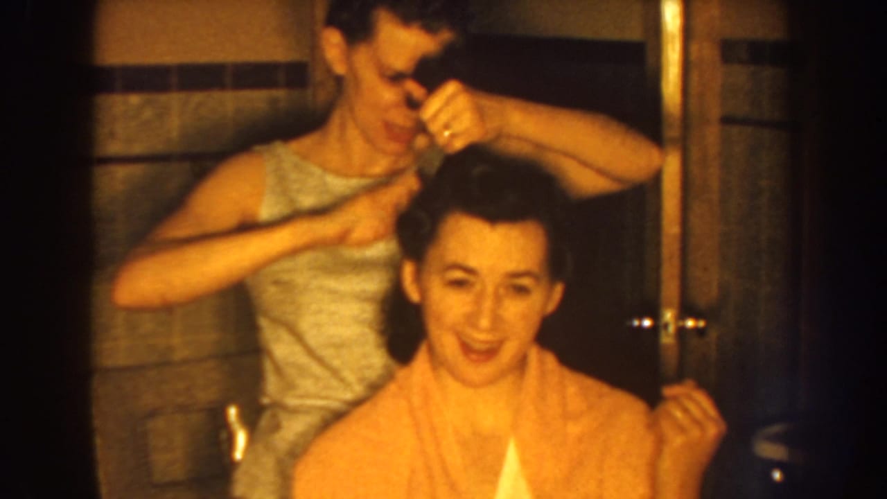Woman reaches across a seated woman's hair while grooming it in a bathroom.