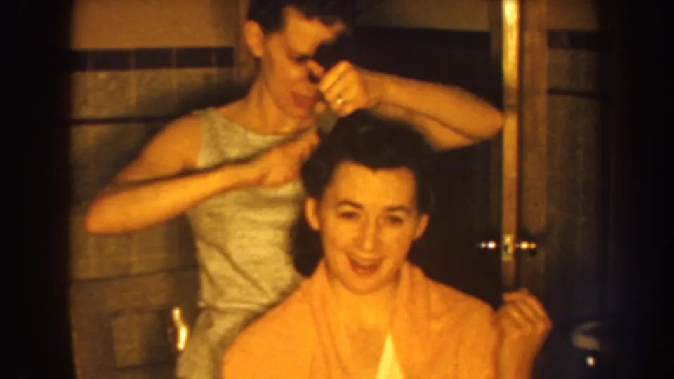 The standing woman works over the seated woman's dark hair in a Nebraska bathroom scene dated to 1959 by clip metadata.