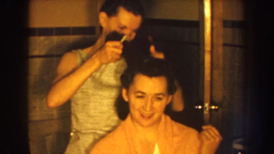 A smiling seated woman wrapped in a yellow towel has her dark hair lifted and arranged in this 1959 Nebraska home movie frame.